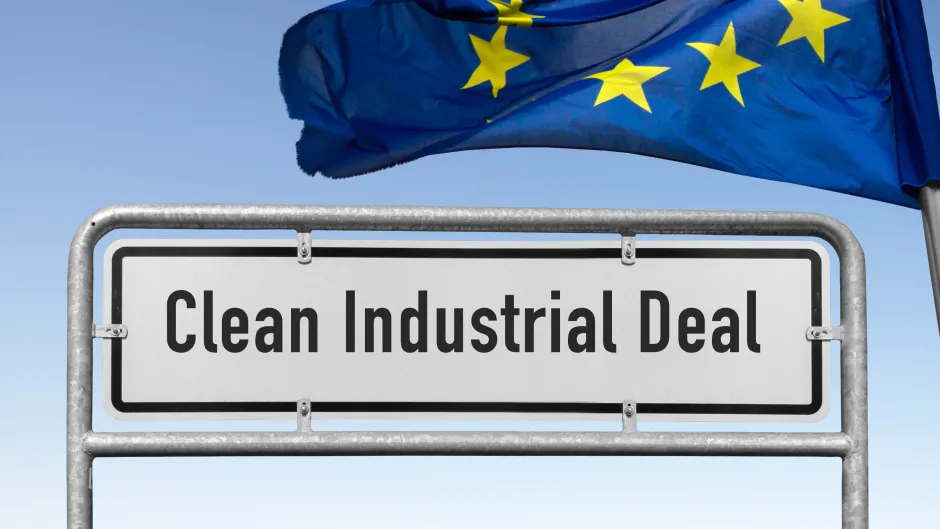 Blick auf ein weißes Ortsangabenschild mit der Aufschrift "Clean Industrial Deal" - daneben weht eine EU-Flagge im Wind vor blauem Hintergrund